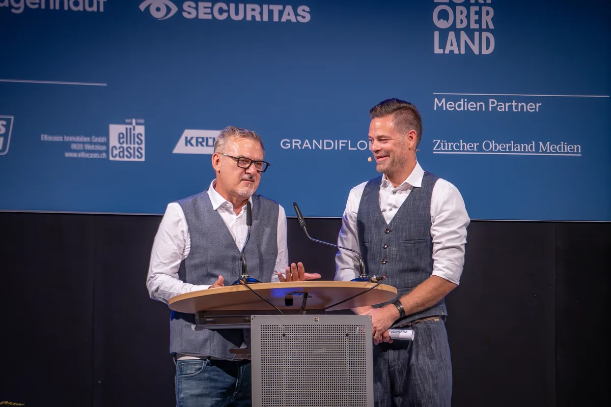 Andreas Künzli und Lukas Studer stehen auf der Bühne vor einer blauen Sponsorenwand. Sie tragen beide ein helles Hemd und eine dunkle Weste.